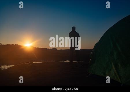 Hinterleuchteter Mann, der mit Sonnenaufgang über dem Bergschatten und einem ruhigen See mit Spiegelung am Morgen steht Stockfoto