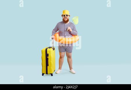 Lustiger fetter Mann in Badeanzug, Hut, Brille und aufblasbarem Ring, der mit seinem Koffer im Studio steht Stockfoto