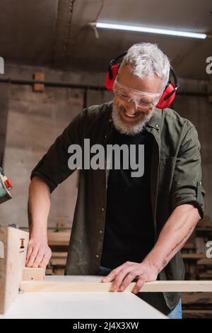 Lächelnder Zimmermann in einer Brille, der in der Werkstatt an einer Jointer-Maschine arbeitet Stockfoto