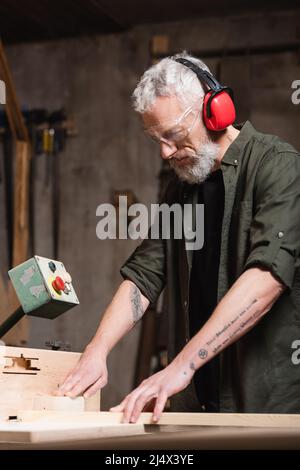 Bärtiger Zimmermann in Ohrenschützern, die an der Jointer-Maschine arbeiten Stockfoto