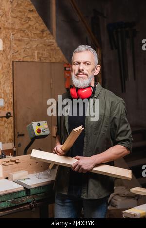 Bärtiger Zimmermann mit Holzdetails und Blick auf die Kamera in der Werkstatt Stockfoto