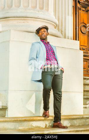 Mann lehnt sich zurück. Modische Newboy Mütze tragen, in hellgrauem Blazer kleiden, rosa gemustert, schwarz unter dem Hemd, schwarze Hose, braune Lederschuhe, Stockfoto