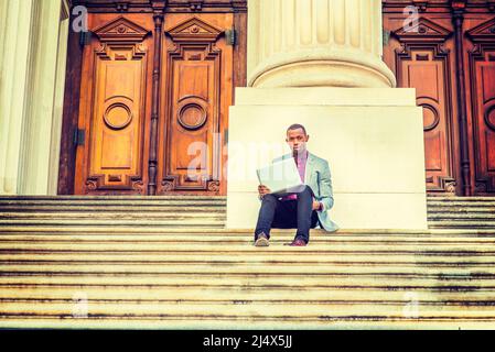 Arbeiten im Freien. Ein junger Schwarzer sitzt auf einer Treppe vor dem Büro im Vintage-Stil und kleidet sich in hellgrauen Blazer, schwarze Hosen, braune Lederschuhe Stockfoto