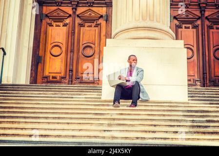 Arbeiten im Freien. Ein junger Schwarzer sitzt auf einer Treppe vor dem Büro im Vintage-Stil und kleidet sich in hellgrauen Blazer, schwarze Hosen, braune Lederschuhe Stockfoto