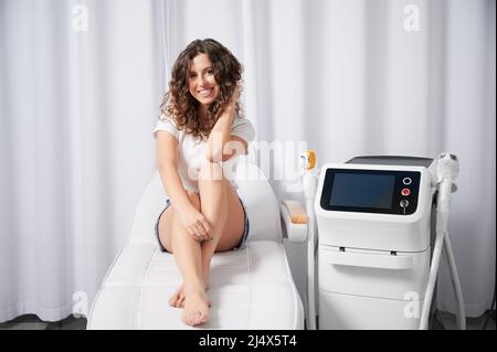 Fröhliche Patientin wartet auf Epilation in der Kosmetologie-Klinik. Glückliche junge Frau, die die Kamera anschaut und lächelt, während sie in der Nähe der Diodenlaser-Haarentfernungsmaschine sitzt. Stockfoto