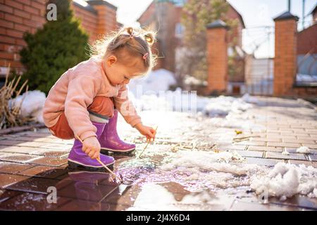 Mädchen spielt in einer Pfütze mit Holzstock am Frühlingstag bei Sonnenlicht Stockfoto