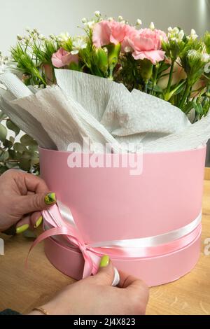 Nahaufnahme der Hände einer Floristin, die weiße und rosafarbene Bänder auf einer pinken Schachtel mit einem Blumenarrangement bindet. Vertikales Foto Stockfoto