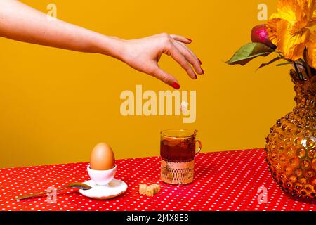 Serviert Frühstückstisch. Food Pop Art Fotografie. Tee, Ei und Blumen auf roter Tischdecke auf gelbem Hintergrund. Interieur im Retro-Stil Stockfoto