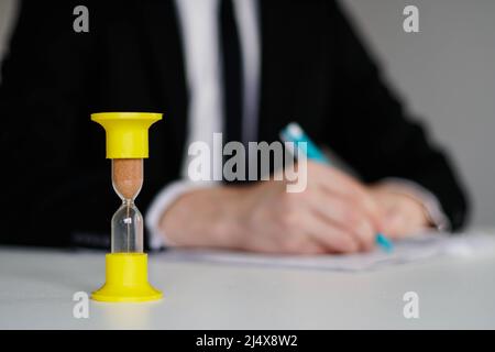 Nahaufnahme von Sanduhr vor dem Geschäftsmann, der mit Dokumenten im Büro arbeitet. Stockfoto