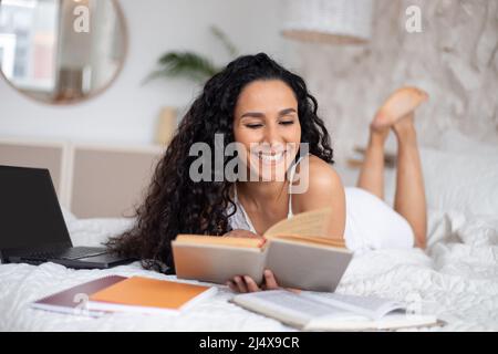 Fröhliche junge kaukasische Brünette Frau mit langen lockigen Haaren im Pyjama liest Bücher und arbeitet mit Laptop Stockfoto