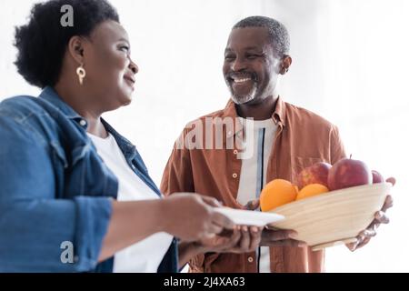 Glücklicher älterer afroamerikanischer Mann, der eine Schüssel mit frischen Früchten in der Nähe einer lächelnden Frau hielt Stockfoto