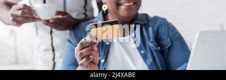 Beschnittene Ansicht einer älteren afroamerikanischen Frau, die ihre Kreditkarte in der Nähe des Laptops hält, und eines verschwommenen Ehemanns mit Tasse und Banner Stockfoto