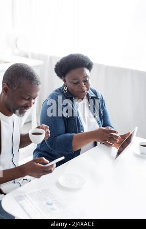 afroamerikanische Frau, die ein digitales Tablet in der Nähe eines fröhlichen Mannes mit einer Tasse Kaffee mit dem Smartphone hält Stockfoto