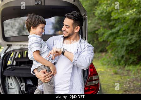 Glücklicher arabischer Mann, der Sohn in den Armen hält und im Sommer mit ihm in der Nähe des Autos mit offenem Kofferraum spielt Stockfoto