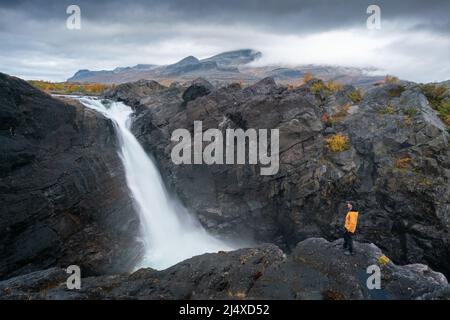 Mann, der auf einem Felsen mit einem mächtigen Wasserfall unter einem dramatischen Himmel steht. Stuor Muorkke Wasserfall im Stora Sjofallet Nationalpark, Schweden. Abenteuer in der Arktis w Stockfoto