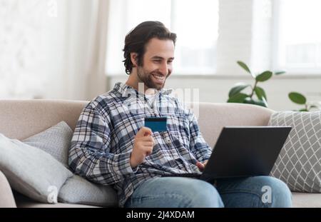 Glücklicher junger europäischer männlicher Shopaholic mit Stoppeln im Hemd zeigt Kreditkarte und schaut auf Laptop sitzt auf dem Sofa Stockfoto