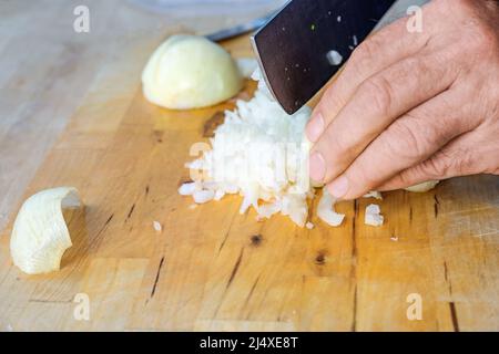 Hand eines Mannes, der Zwiebeln mit einem Küchenmesser auf einem hölzernen Schneidebrett in kleine Würfel schneidet, Platz kopieren, ausgewählter Fokus, enge Schärfentiefe Stockfoto