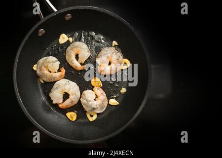 Frische Garnelen werden mit Knoblauch in Olivenöl in einer Pfanne auf dem schwarzen Herd anbraten, Vorbereitung für eine leckere Vorspeise, Kopierraum, hoher Winkel Stockfoto