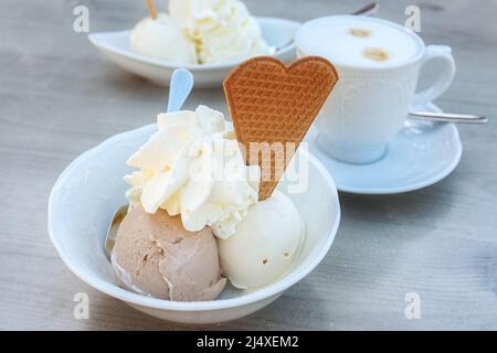Eisdessert mit Haselnuss- und Vanilleschaufeln, Schlagsahne und Keks in einer weißen Schüssel und einer Tasse Kaffee auf einem grauen Tisch im Freien auf einer Straße Stockfoto