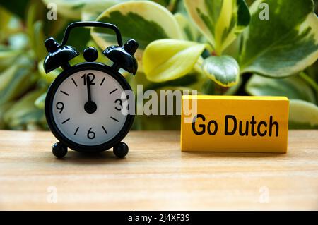 Wecker zeigt auf 12 Uhr mit Go Dutch Text auf gelbem Holzblock. Mittagskonzept Stockfoto