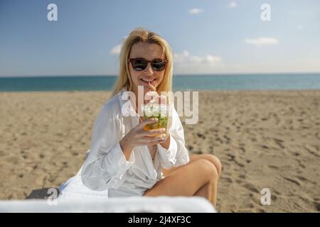 Porträt einer Dame, die einen Cocktail trinkt und die Kamera anlächelt, während sie am leeren Strand sonnenbaden Stockfoto