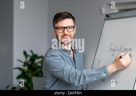 Selbstbewusster reifer männlicher Lehrer, der Online-Unterricht gibt, neues Material erklärt, englische Regeln auf Tafel schreibt Stockfoto