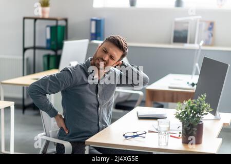 Reifer Geschäftsmann massiert Nacken- und Rückenschmerzen, leidet nach der Computerarbeit unter Schmerzen, sitzt am Arbeitsplatz Stockfoto