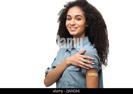 Heitere geimpfte afroamerikanische Frau, die Arm mit medizinischem Pflaster zeigt und lacht, Frau erhält Impfdosis gegen Covid, Gips auf der Schulter, isoliert auf Weiß. Gesundheitskonzept Stockfoto