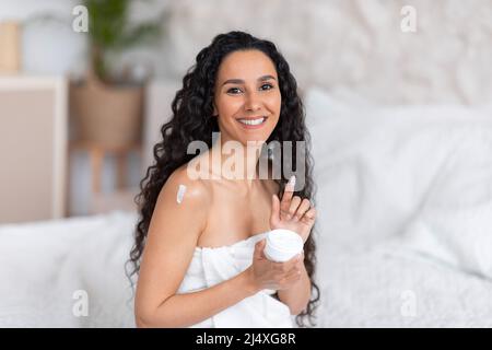 Fröhliche tausendjährige kaukasische Brünette weiblich mit langen lockigen Haaren im Handtuch gilt Creme auf der Schulter, halten Glas Stockfoto