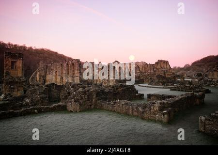 Die Wand des wärmenden Hauses, hinter dem das Refektorium der zerstörten Rievaulx Zisterzienserabtei bei Sonnenaufgang und nach dem Mond aufgegangen ist Stockfoto