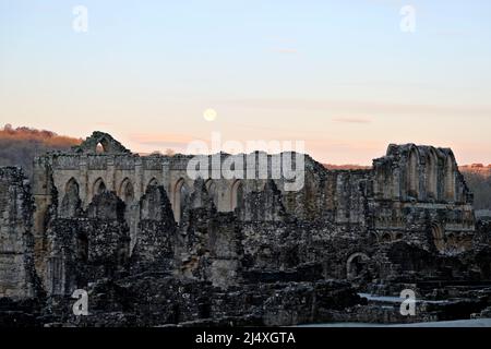 Die Wand des wärmenden Hauses, hinter dem das Refektorium der zerstörten Rievaulx Zisterzienserabtei bei Sonnenaufgang und nach dem Mond aufgegangen ist Stockfoto