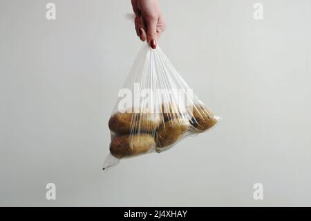 Die Hand der Frau hält Kartoffeln in einem Plastikbeutel. Lebensmittel in Zellophanverpackung auf grauem Hintergrund verpackt. Nicht ökologische Verpackungen Stockfoto