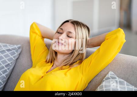 Glückliche junge europäische blonde Frau in gelben Kleidern mit geschlossenen Augen, die im Wohnzimmer auf dem Sofa ruht Stockfoto