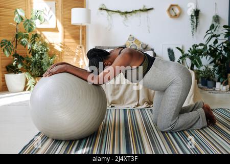 Afrikanische Schwangere, die auf den Knien steht und sich auf den Fitnessball lehnt, fühlt während der Wehen Schmerzen und gebiert zu Hause Stockfoto