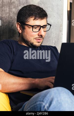 Ein hübscher bärtiger Mann in einer Brille blickt in einem modernen Büro vor dem Hintergrund einer Betonwand auf seinen Laptop-Bildschirm Stockfoto