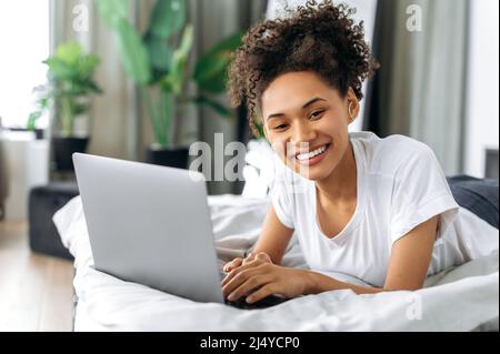 Schöne positive afroamerikanische lockige Brünette liegt zu Hause auf einem Bett auf dem Bauch, nutzt einen Laptop, durchsucht das Internet, chattet in sozialen Netzwerken, schaut in die Kamera, lächelt glücklich Stockfoto