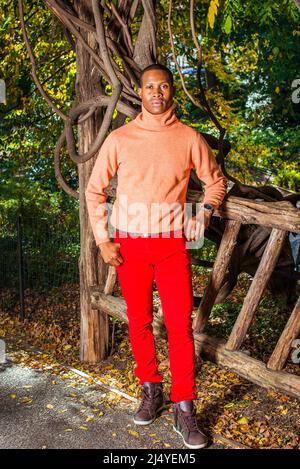 Männer Herbst Casual Fashion. Kleid in hellorange Pullover mit hohem Kragen, rote Hosen, gemusterte Stiefel Schuhe, Tragen Armbanduhr, ein junger schwarzer Kerl i Stockfoto