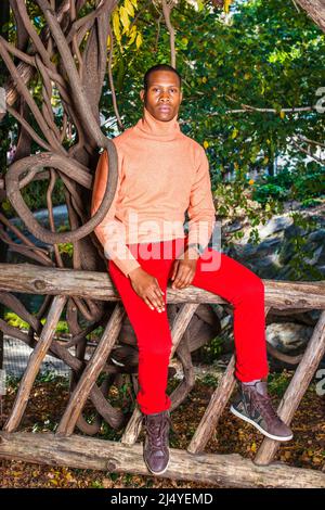 Männer Herbst Casual Fashion. Kleid in hellorange Pullover mit hohem Kragen, rote Hosen, gemusterte Stiefel Schuhe, Tragen Armbanduhr, ein junger schwarzer Kerl i Stockfoto