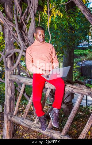 Mann, Der Draußen Arbeitet. In hellorangefarbenem Pullover mit hohem Kragen, roten Hosen, gemusterten Stiefelschuhen, sitzt ein junger Schwarzer auf einem Holzzaun, Stockfoto