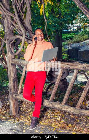 Mann, Der Draußen Arbeitet. In hellorangenen Pullover mit hohem Kragen, roten Hosen, gemusterten Stiefelschuhen kleidet, steht ein junger Schwarzer am Zaun, Smili Stockfoto