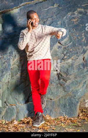 Warten. Dressing in Creme, gemusterter, kragenloser Pullover, rote Jeans, Stiefelschuhe, Ein junger Schwarzer steht in der Armbanduhr am Felsen, lächelt, Stockfoto