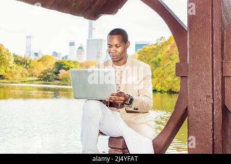 Geschäftsmann, Der Arbeitet. Ein junger Schwarzer sitzt in einem Big an einem See und trägt einen cremefarbenen Blazer, einen kragenlosen Pullover, eine weiße Hose und eine Armbanduhr Stockfoto