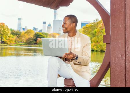 Geschäftsmann, Der Arbeitet. Ein junger Schwarzer sitzt in einem großen Cit am See und trägt einen cremefarbenen Blazer, einen kragenlosen Pullover, eine weiße Hose und eine Armbanduhr Stockfoto