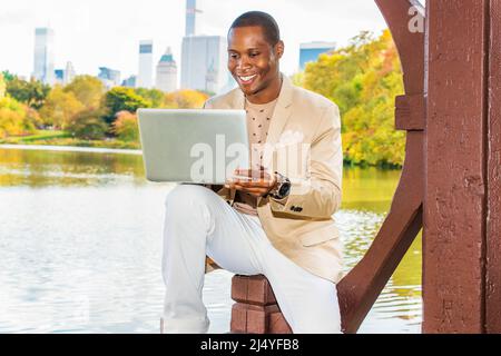 Geschäftsmann, Der Arbeitet. Ein junger Schwarzer sitzt in einem Big an einem See und trägt einen cremefarbenen Blazer, einen kragenlosen Pullover, eine weiße Hose und eine Armbanduhr Stockfoto
