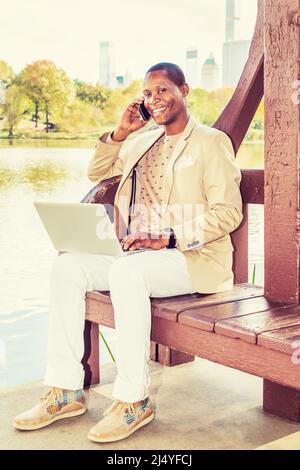 Geschäftsmann, Der Arbeitet. Ein junger Schwarzer sitzt an einem See in der Großstadt und kleidet sich in einen cremefarbenen Blazer, einen kragenlosen Pullover, weiße Hosen, Turnschuhe Stockfoto