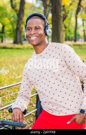 Mann Liebt Musik. In gemustertem kragenlosem Pullover, roter Hose, Kopfhörer tragend, kippt ein junger Schwarzer auf einem Fahrrad im Park, lächelnd, Stockfoto