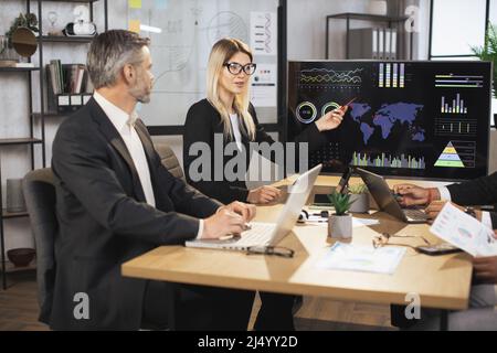 Vorstand verschiedener multiethnischer Direktoren Geschäftsleute bei jährlichen Treffen im Bürozimmer, hören reife blonde Dame zu und diskutieren Arbeitsergebnisse. TV-Bildschirm mit Karte und Karten im Hintergrund. Stockfoto