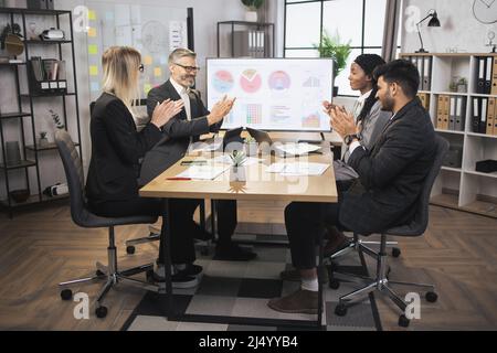 Gute Ergebnisse, Geschäft, Gratulation und Wertschätzung Konzept. Zwei zufriedene, vielseitige Geschäftsleute in formeller Kleidung, die im Konferenzsaal am Tisch sitzen und applaudieren. Konzentrieren Sie sich auf den jungen Mann Stockfoto