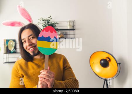 Junge kaukasische Mann trägt gefälschte Kaninchenohren hält eine bunte Papierfigur eines Ostereiers auf einem Stock Ostern Konzept Medium closeup kopieren Raum Hause Hintergrund . Hochwertige Fotos Stockfoto