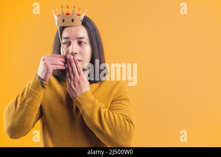 Attraktiver junger Mann, der Papierkrone über seinem Kopf hält und seine Lippen berührt, schaut weg, isoliert, Studio, gelber Hintergrund, Kopierraum. Hochwertige Fotos Stockfoto